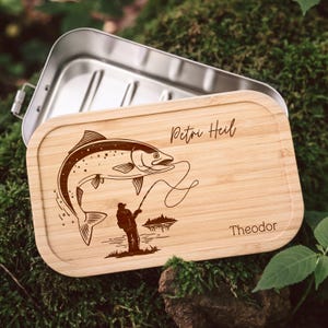 Op de afbeelding: Een bamboe lunchbox met een roestvrijstalen binnenkant. De houten deksel heeft een gegraveerde illustratie van een vis, een visser en de teksten "Petri Heil" en "Theodor". De deksel is lichtbruin.