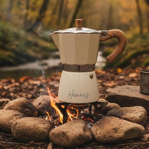 Può includere: Una caffettiera color crema con manico e pomello in legno, fumante sopra un piccolo fuoco da campo. Il nome "Hannes" è scritto sul davanti. La caffettiera è posta su un fornello portatile circondato da rocce, con uno sfondo sfocato di foresta.
