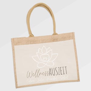 Puede incluir: Bolso tote de yute beige con un panel frontal crema. El panel presenta un dise&ntilde;o de flor de loto blanca y las palabras "Wellness Auszeit". El bolso tiene asas beige a juego.