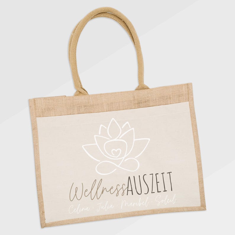 Puede incluir: Bolso tote de yute beige con un panel frontal crema. El panel presenta un dise&ntilde;o de flor de loto blanca y las palabras "Wellness Auszeit". El bolso tiene asas beige a juego.