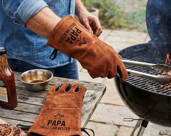 May include: Brown leather grilling gloves with the text "PAPA GRILLT AM BESTEN" and a grilling graphic. One glove is on a person's hand, and another is on a wooden table. A grill and steak are in the background.