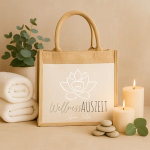 Puede incluir: Un bolso tote beige con un panel blanco con un logotipo de flor de loto y el texto "Wellness AUSZEIT". El bolso tiene una textura de yute y asas resistentes. Dos velas encendidas, piedras lisas y toallas enrolladas en el fondo.