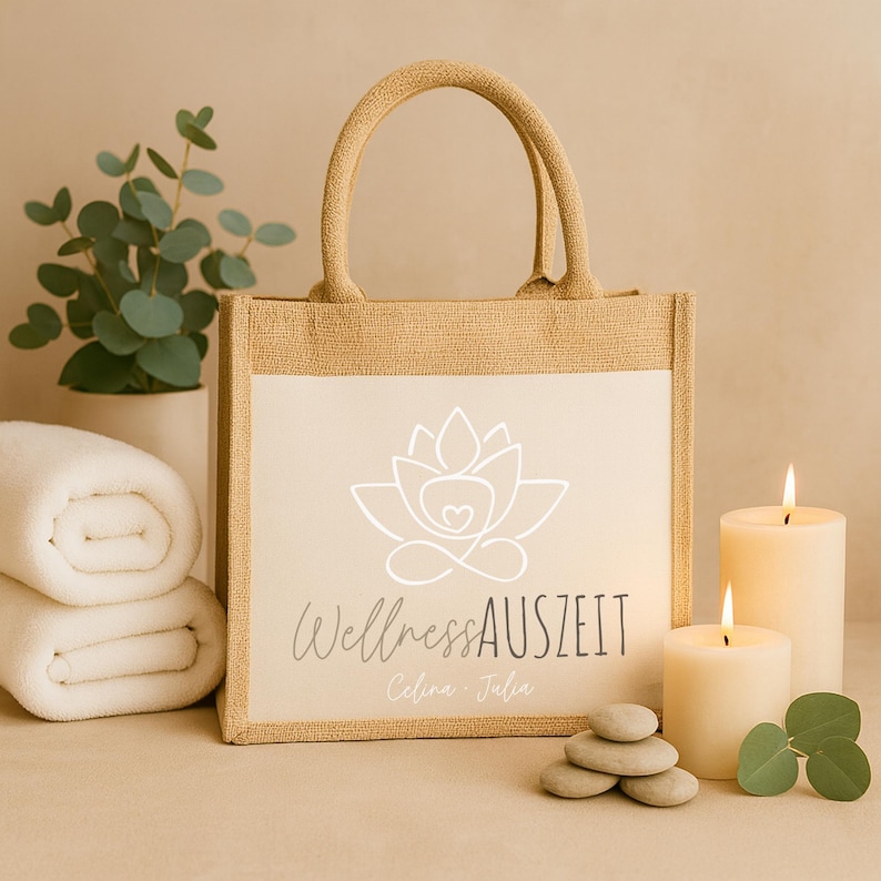 Puede incluir: Un bolso tote beige con un panel blanco con un logotipo de flor de loto y el texto "Wellness AUSZEIT". El bolso tiene una textura de yute y asas resistentes. Dos velas encendidas, piedras lisas y toallas enrolladas en el fondo.