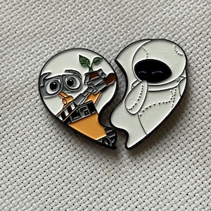 Könnte beinhalten: Herzförmiger Emaille-Pin mit Charakteren aus dem Film Wall-E. Eine Seite zeigt Wall-E mit einer Pflanze, die andere Eve. Der Pin hat einen schwarzen Rand und ist vor einem strukturierten, hellgrauen Hintergrund platziert.