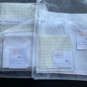 May include: Three white Aida cloth samples in clear plastic bags. The bags are labeled with the text "Order" and the type of cloth, including the size in inches. The cloth is 14 count Aida, 10 count Aida, and 28 count Aida.