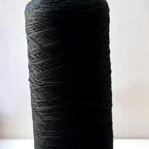May include: A tall, cylindrical spool of solid black yarn. The thread is tightly wound in a crisscross pattern around the central core, creating a uniform, textured surface that extends from the top to the bottom of the spool.
