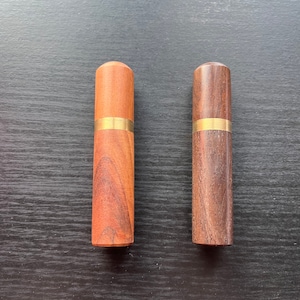 May include: Two wooden needle cases with a gold band around the middle. The case on the left is a reddish brown colour and the case on the right is a dark brown colour.