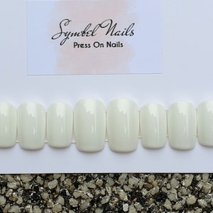 Könnte beinhalten: Ein Set mit zehn weißen, aufklebbaren Nägeln in Sargform. Die Nägel sind in einer Reihe auf weißem Hintergrund angeordnet. Der Text "Symbol Nails Press On Nails" ist oben im Bild sichtbar.