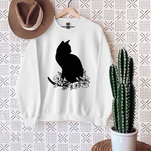 May include: White sweatshirt with a black silhouette of a cat sitting with flowers. The sweatshirt is hanging on a wooden hanger against a white background with a black and white pattern.