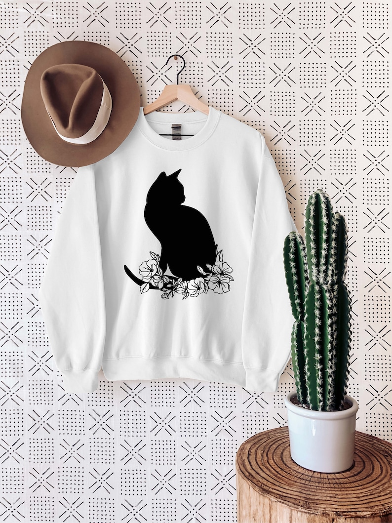 May include: White sweatshirt with a black silhouette of a cat sitting with flowers. The sweatshirt is hanging on a wooden hanger against a white background with a black and white pattern.