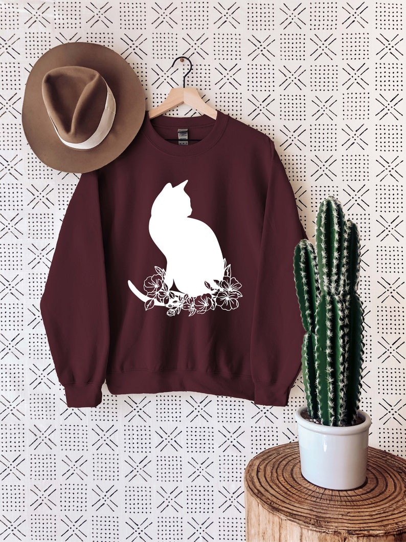May include: A maroon sweatshirt with a white silhouette of a cat sitting with flowers. The sweatshirt is hanging on a hanger against a white background with a black and white geometric pattern.