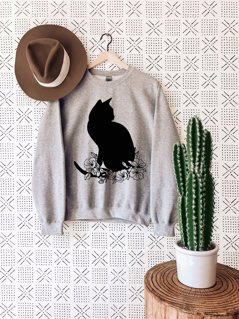 May include: A gray sweatshirt with a black silhouette of a cat sitting on a branch with flowers. The sweatshirt is hanging on a hanger against a white background with a black geometric pattern.