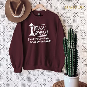 May include: Maroon sweatshirt with white text that reads "Black Queen Most Powerful Piece in the Game". The sweatshirt features a white chess queen graphic.