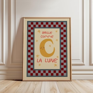 Puede incluir: Impresión artística enmarcada con una ilustración de una luna creciente amarilla sonriente, rodeada de estrellas y el texto francés "Brille comme la Lune" en rojo. La obra está enmarcada con un borde a cuadros rojo y gris.