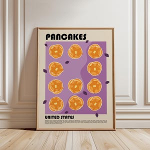 May include: A framed wall art print featuring the word PANCAKES at the top and UNITED STATES at the bottom. The center displays eleven orange pancake illustrations with butter pats on a purple background, accented by small dark purple leaf shapes.