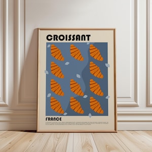 May include: A framed poster featuring the word CROISSANT at the top and FRANCE at the bottom. The center displays a blue rectangular field with twelve orange croissant illustrations and small light blue leaf shapes. Text at the bottom describes the pastry.