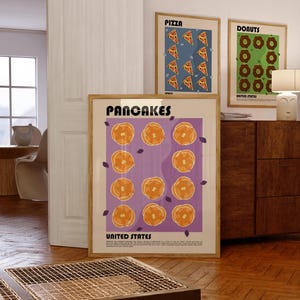 May include: A framed wall art print titled PANCAKES UNITED STATES featuring a grid of eleven golden-brown pancakes on a purple background, with small dark purple leaf accents and descriptive text at the bottom, displayed in a light wood frame.