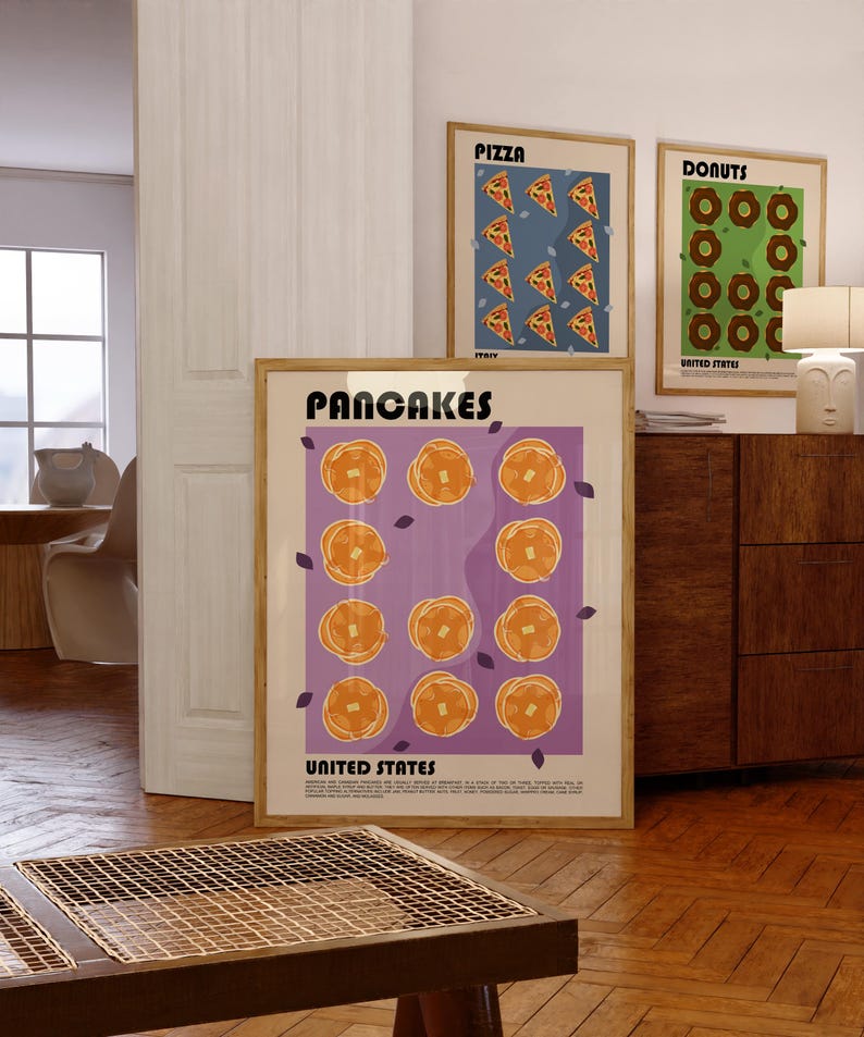 May include: A framed wall art print titled PANCAKES UNITED STATES featuring a grid of eleven golden-brown pancakes on a purple background, with small dark purple leaf accents and descriptive text at the bottom, displayed in a light wood frame.