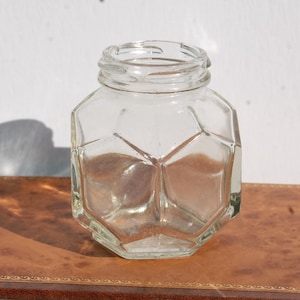 May include: A clear glass jar with a distinct hexagonal geometric shape and a threaded opening at the top. The container features flat, angled panels that create a multifaceted appearance, reflecting light across its transparent, smooth surface.