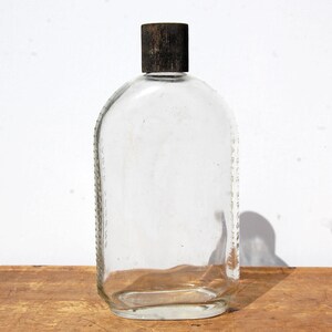 May include: Clear glass bottle with a rounded shape and a dark brown cap. The bottle has a rectangular body with rounded edges and a textured cap. The bottle is empty and sits on a wooden surface.