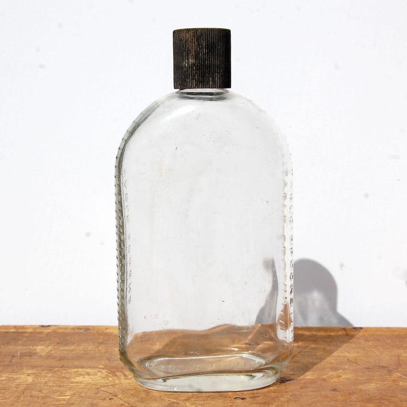 May include: Clear glass bottle with a rounded shape and a dark brown cap. The bottle has a rectangular body with rounded edges and a textured cap. The bottle is empty and sits on a wooden surface.