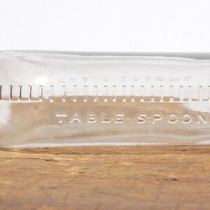 May include: A clear glass bottle with a dark brown cap. The bottle is marked with measurements in tablespoons. The words "TABLE SPOONS" are etched into the glass. The bottle is lying horizontally on a wooden surface.
