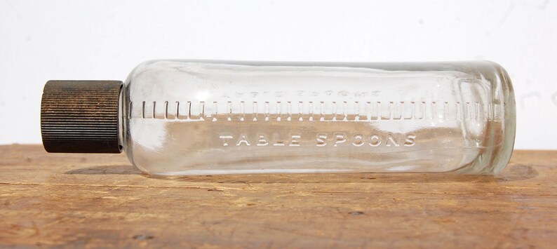 May include: A clear glass bottle with a dark brown cap. The bottle is marked with measurements in tablespoons. The words "TABLE SPOONS" are etched into the glass. The bottle is lying horizontally on a wooden surface.