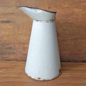 Puede incluir: Una jarra de esmalte blanco roto vintage con un pico. La jarra tiene un cuerpo cónico y un borde ligeramente oxidado, lo que sugiere edad y uso. El fondo es una superficie de madera.