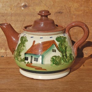 May include: A ceramic teapot with a brown lid, handle, and spout. The body is cream-coloured with a hand-painted scene of a house with a brown roof, green trees, and a path. A row of small blue dots circles the top edge of the teapot body.