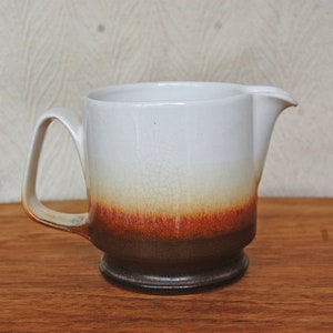 May include: A white ceramic creamer with a brown and orange glaze. The creamer has a handle and a spout.