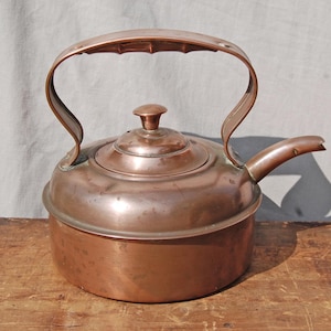 Puede incluir: Una tetera de cobre vintage con un acabado desgastado y un asa curva. La tetera tiene una tapa con un pomo en la parte superior y un pico en el lateral.