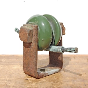 May include: An industrial-style object featuring two green, circular insulators held within a rusty, U-shaped metal bracket. A copper wire is threaded through the insulators. The object is set against a light background and a wooden surface.