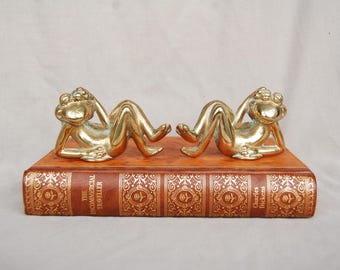 May include: Two brass frog figurines in a relaxed pose, resting on a vintage book. The frogs are golden and detailed, with their arms supporting their heads. The book is brown with gold embossed lettering and designs.