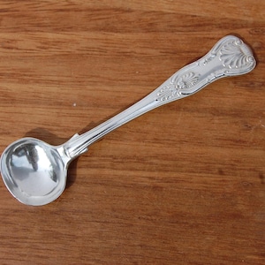 Puede incluir: Una pequeña cuchara plateada ornamentada, con un cuenco redondeado y un mango decorativo. La cuchara se encuentra sobre un fondo de madera cálida, resaltando su superficie pulida y su intrincado diseño. Esta cuchara de estilo antiguo es probablemente para servir o exhibir.