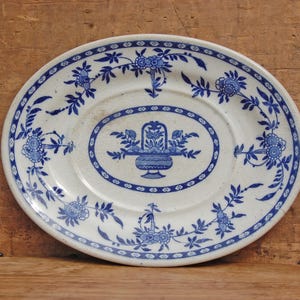 May include: Oval ceramic plate with a white base and blue floral design. The plate features a central oval with a floral basket motif and a border of flowers and leaves. The plate is set against a wooden surface.