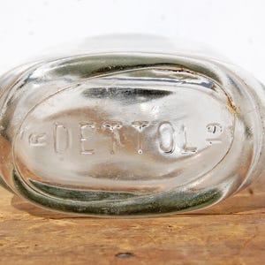 May include: Clear glass bottle with the embossed word "DETTOL". The bottle has an oval shape and is resting on a wooden surface. The glass has a slight green tint around the edges.