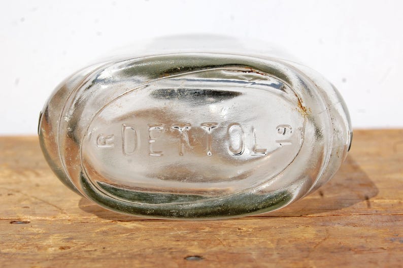 May include: Clear glass bottle with the embossed word "DETTOL". The bottle has an oval shape and is resting on a wooden surface. The glass has a slight green tint around the edges.