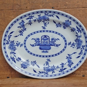 Può includere: Piatto ovale in ceramica bianca con un motivo floreale blu. Il piatto presenta un ovale centrale con un cesto di fiori e un bordo di fiori e foglie. Il bordo ha un motivo floreale blu. Il piatto è su una superficie di legno.