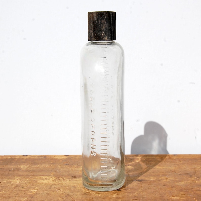 May include: A clear glass bottle with a dark brown cap. The bottle has measurement markings in tablespoons and a scale along the side. The bottle is cylindrical and sits on a wooden surface.