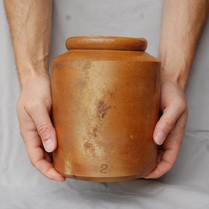 Puede incluir: Un tarro cilíndrico de gres marrón, con una parte superior redondeada y una base ligeramente más ancha. El tarro tiene el número "2" estampado cerca de la parte inferior y muestra signos de edad con algo de decoloración. El tarro está sostenido por dos manos.