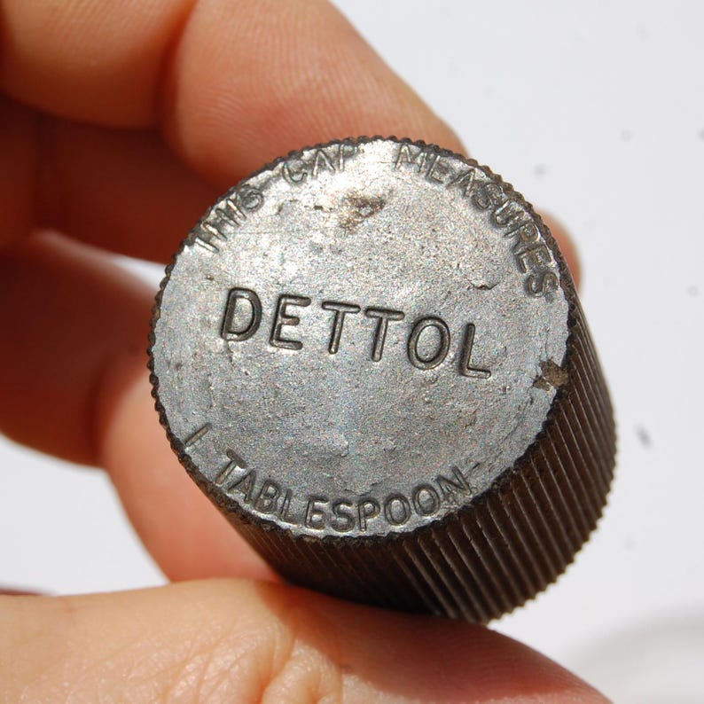 May include: Close-up of a vintage Dettol bottle cap. The cap is silver with the word "DETTOL" in the center. The text "THIS CAP MEASURES" and "1 TABLESPOON" are also visible. The cap has a ridged, brown metal side.