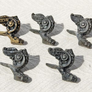 May include: Five metal furniture feet, each featuring a detailed scrollwork design and a claw-shaped base. Four of the feet have a dark, weathered finish, while one foot displays a worn, metallic gold-toned patina with visible dark accents.