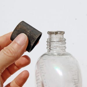 May include: A clear glass bottle with a rounded body and a narrow neck. A dark metal cap with a ridged texture is held near the bottle's opening. The bottle has measurement markings on the side.