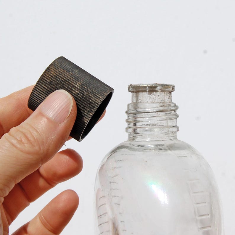 May include: A clear glass bottle with a rounded body and a narrow neck. A dark metal cap with a ridged texture is held near the bottle's opening. The bottle has measurement markings on the side.