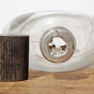 May include: A clear glass flask with a metal neck and a separate, dark brown, ribbed cap. The flask is oval-shaped and lies on a wooden surface. The cap is cylindrical.