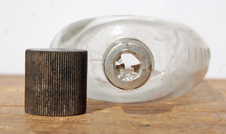 May include: A clear glass flask with a metal neck and a separate, dark brown, ribbed cap. The flask is oval-shaped and lies on a wooden surface. The cap is cylindrical.
