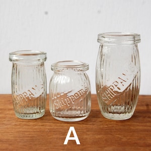 May include: Three clear glass jars of varying sizes, each featuring vertical ribbed textures and the embossed text Shippam's on the side. The jars have a wide mouth opening and a thick rim, standing upright in a row. The letter A is printed below.