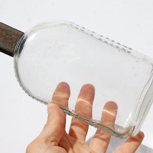 May include: A clear, rectangular glass bottle with a rounded top and a dark brown, ribbed cap. The bottle has a textured edge and is held in a hand.