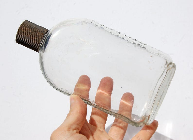 May include: A clear, rectangular glass bottle with a rounded top and a dark brown, ribbed cap. The bottle has a textured edge and is held in a hand.
