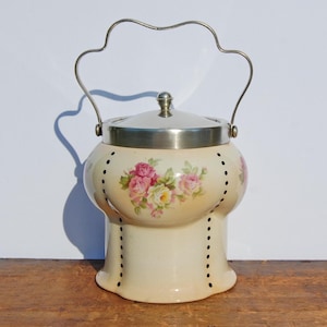 Könnte beinhalten: Eine antike Keramik-Keksdose mit einem silbernen Deckel und Griff. Die Dose ist mit einem Blumenmuster aus rosa Rosen und grünen Blättern auf cremefarbenem Hintergrund verziert.